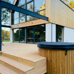 Nowoczesny dom szkieletowy z drewnianym tarasem i jacuzzi na zewnątrz, widok z poziomu gruntu na taras i część elewacji budynku.