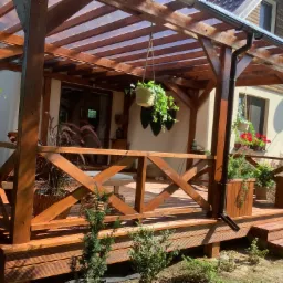 Zadaszona drewniana konstrukcja z elementami dekoracyjnymi, roślinami w donicach i wiszących koszach, z widocznym fragmentem domu w tle.