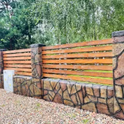 Nowoczesne ogrodzenie z drewnianymi przęsłami i kamienną podmurówką na tle zieleni, widoczna skrzynka elektryczna po lewej stronie.