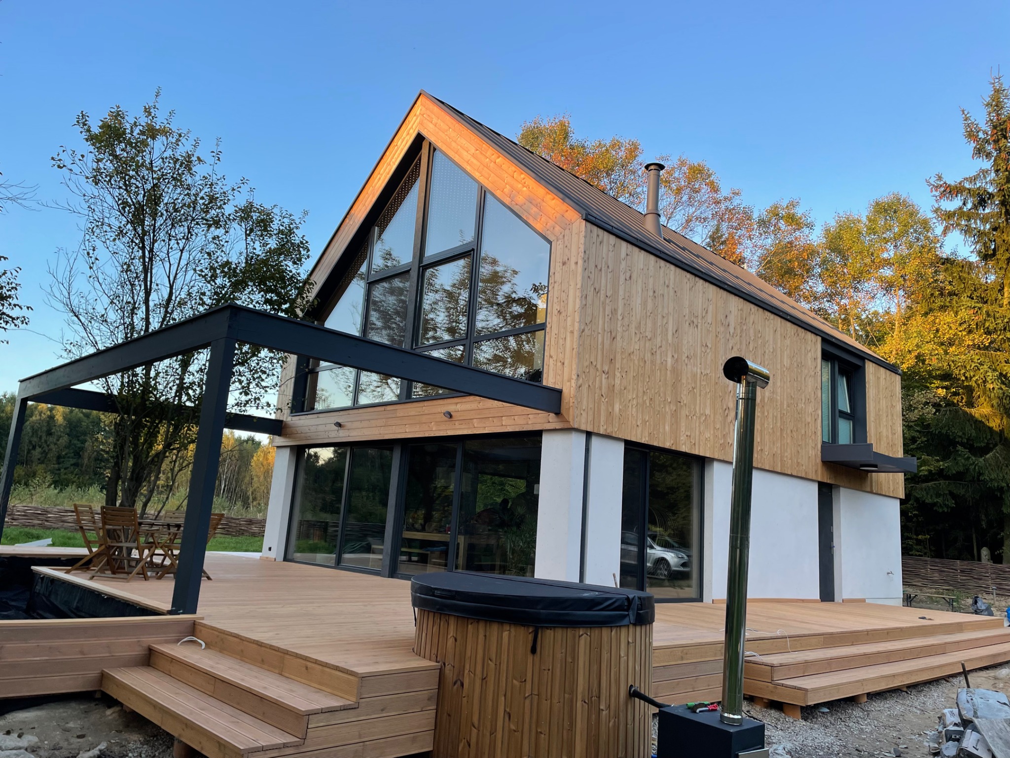 Nowoczesny dom z drewnianą elewacją i dużymi przeszkleniami, taras z drewna z jacuzzi i metalową pergolą, widok na ogród i błękitne niebo.