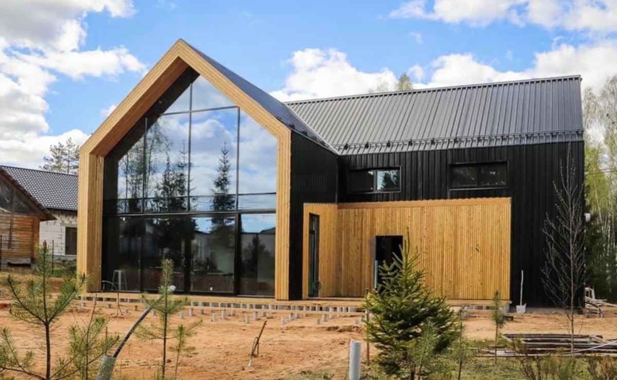 Nowoczesny dom z czarną elewacją i dużym przeszkleniem w kształcie trójkąta, widoczne przygotowania do budowy tarasu na gruncie.