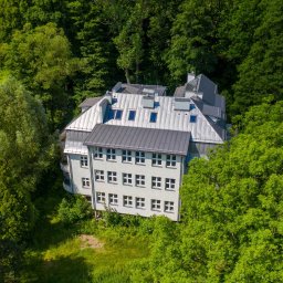 Project Media Piotr Wróbel - Zdjęcie z drona przedstawia odnowiony, trzypiętrowy budynek z szarą, metalową dachówką i białymi oknami, otoczony zielonymi drzewami i trawnikiem.