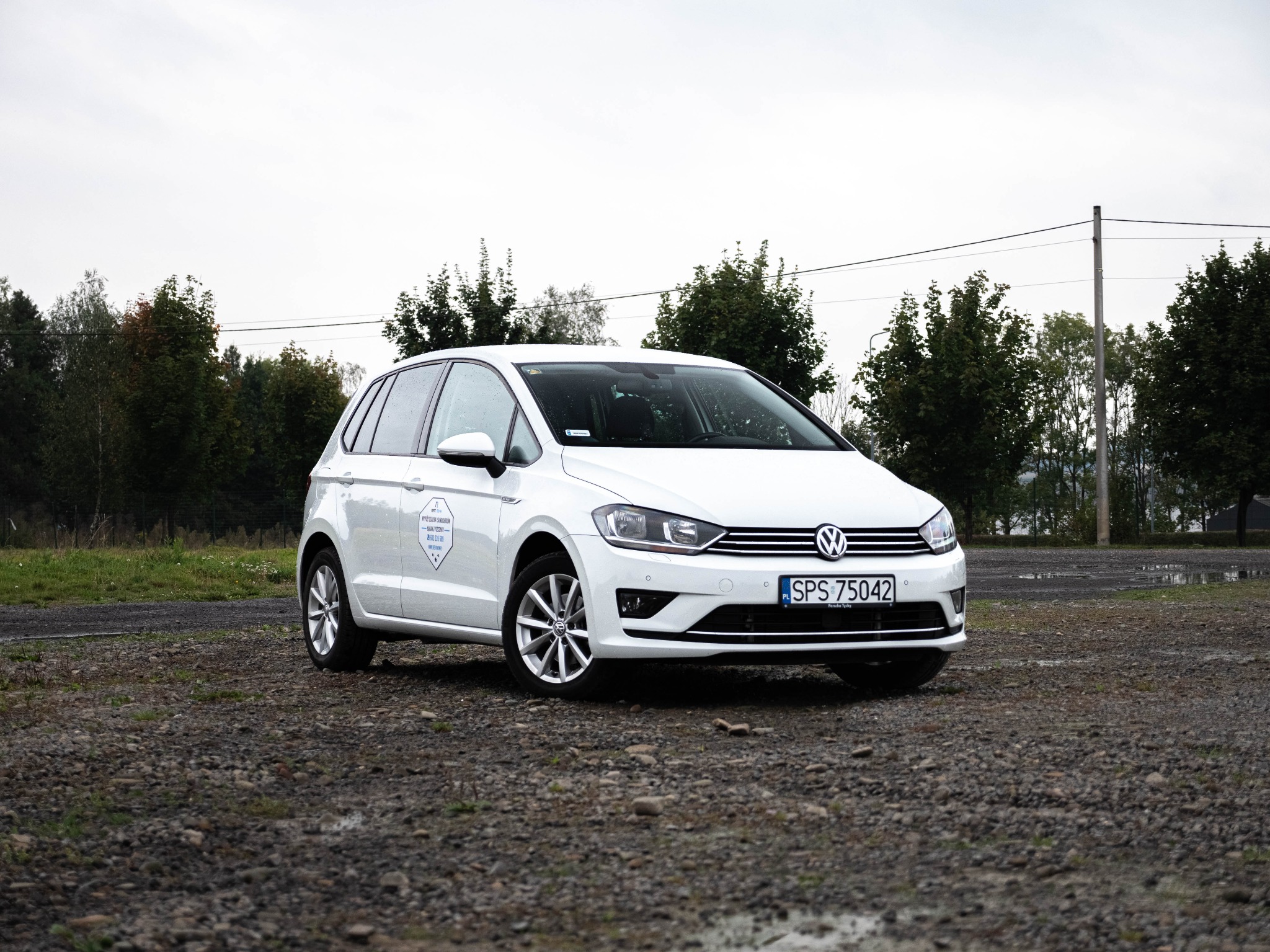 Biały samochód Volkswagen Golf na żwirowym parkingu w pochmurny dzień, z naklejką reklamową na tylnej szybie po stronie kierowcy.