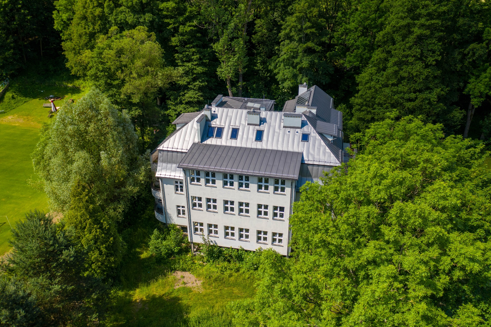 Zdjęcie z drona przedstawia odnowiony, trzypiętrowy budynek z szarą, metalową dachówką i białymi oknami, otoczony zielonymi drzewami i trawnikiem.