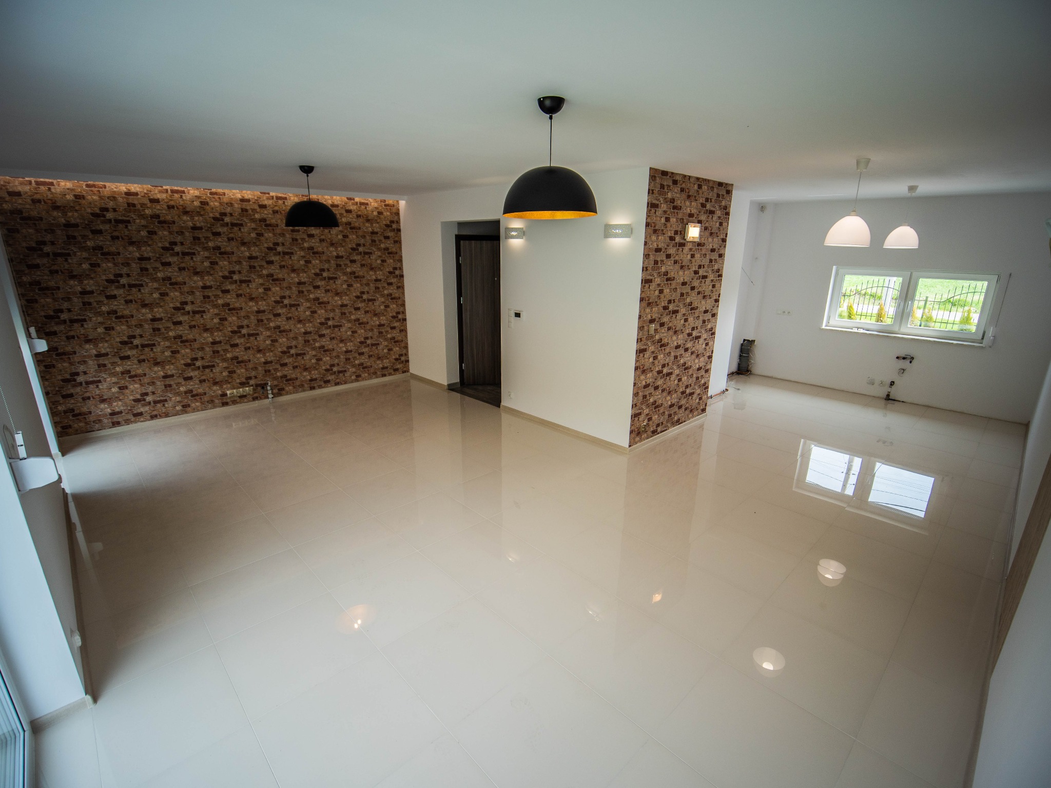 Wykończone wnętrze: białe, błyszczące płytki podłogowe, ściany z dekoracyjną cegłą, nowoczesne lampy wiszące i ścienne, duże okno z widokiem na zieleń.