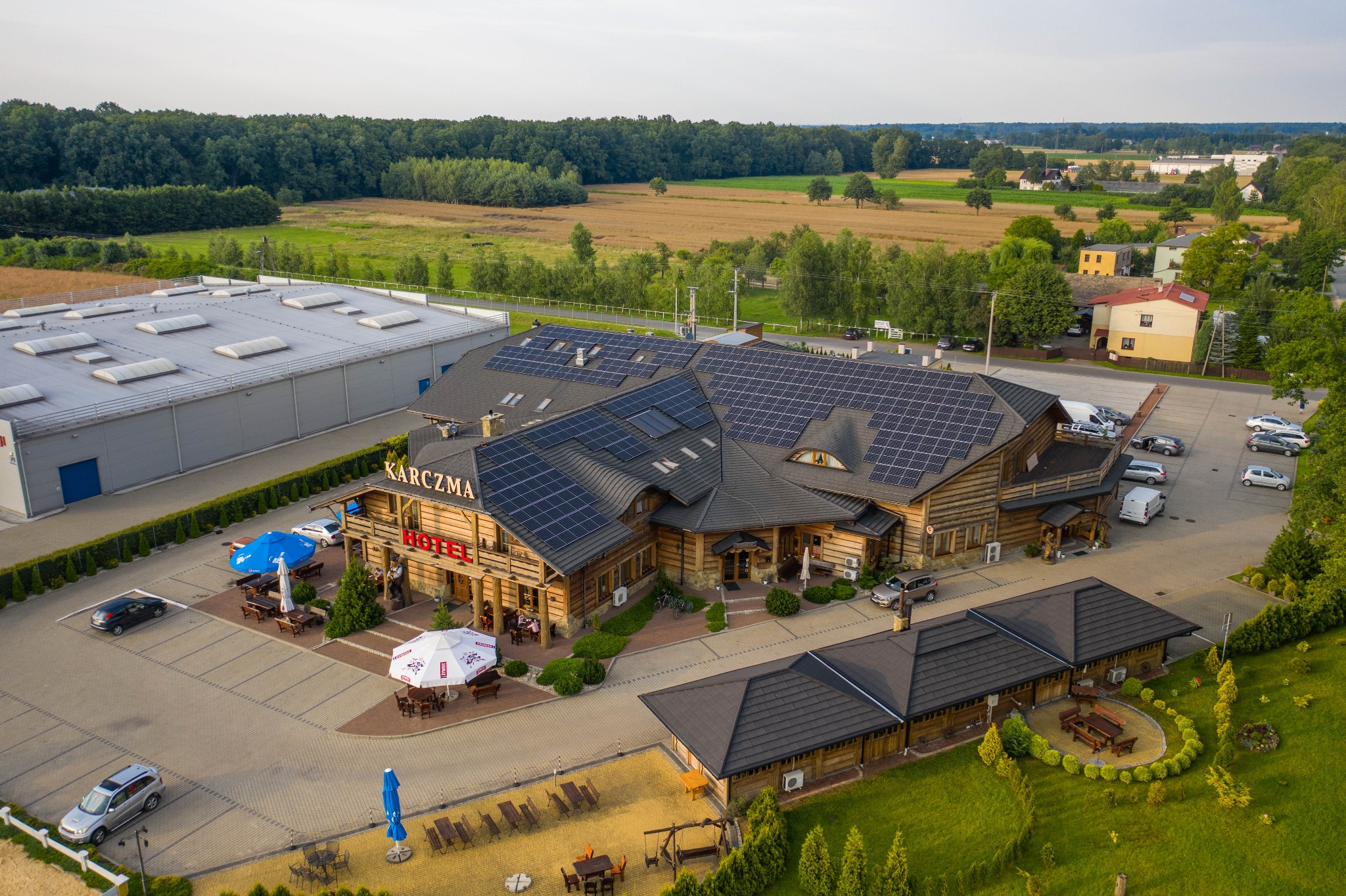 Widok z lotu ptaka na drewnianą karczmę z panelami słonecznymi na dachu, otoczoną parkingiem i zielenią, w tle pola i las.