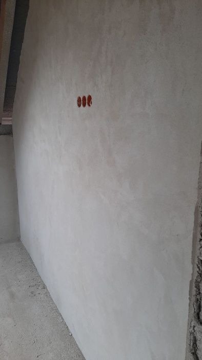 Świeżo położona, jasnoszara warstwa gładzi na ścianie z widocznymi trzema otworami na gniazdka elektryczne, surowy stan wykończenia wnętrza.