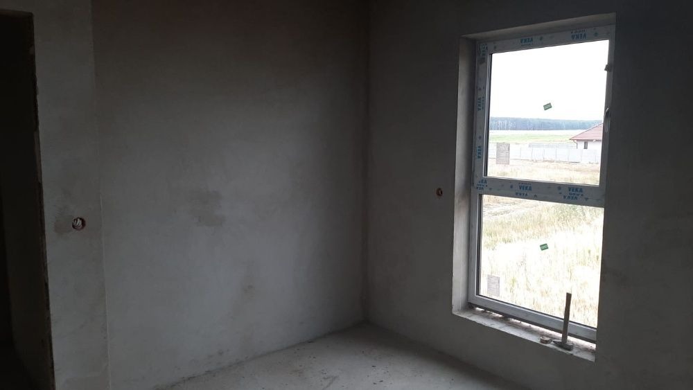 Surowe, szare ściany w nowo wykończonym pomieszczeniu z białym oknem PCV; widoczny fragment podłogi i sufitu, na ścianie widoczne gniazdko elektryczne.