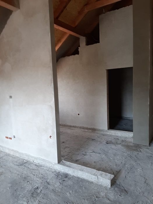 Wnętrze w trakcie prac wykończeniowych: surowe ściany pokryte świeżym tynkiem, widoczne drewniane elementy konstrukcji dachu, betonowa podłoga z widocznymi niedoskonałościami, otwór drzwiowy...