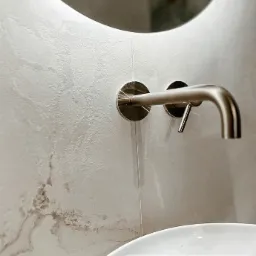 Minimalistyczna łazienka: bateria ścienna w kolorze szczotkowanego niklu, okrągłe lustro z podświetleniem LED i umywalka nablatowa na tle ściany z efektem trawertynu.