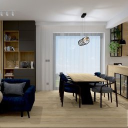 DG Projekty Wnętrz Diana Grabowska - Wnętrze: kobieta siedzi na granatowej sofie w salonie z drewnianą zabudową, regałem i stołem jadalnym. Widok na kuchnię z drewnianymi szafkami i zielonymi roślinami.