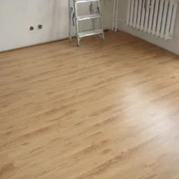 Jasne panele podłogowe ułożone w pokoju z białymi ścianami, widoczny grzejnik i aluminiowa drabina oparta o ścianę.