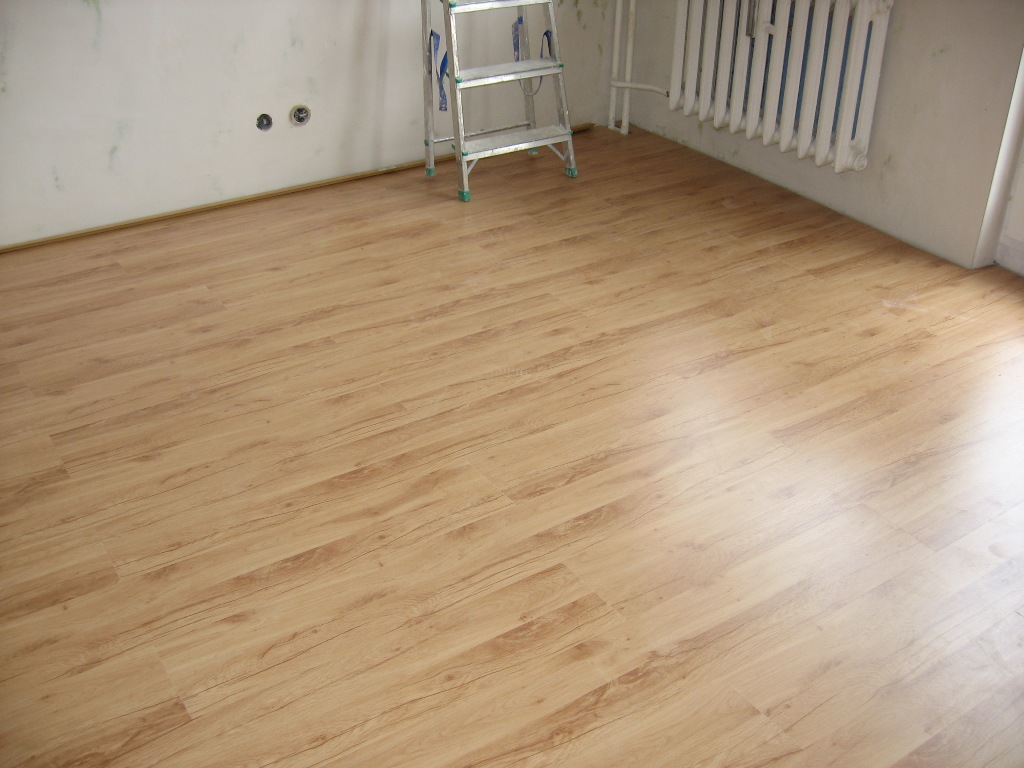 Jasne panele podłogowe ułożone w pokoju z białymi ścianami, widoczny grzejnik i aluminiowa drabina oparta o ścianę.