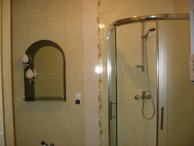 Wykończona łazienka z kabiną prysznicową z chromowanymi elementami i lustrem z oświetleniem w stylu retro, wyłożona jasnymi płytkami z dekoracyjnym pasem z motywem roślinnym.