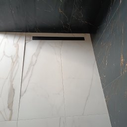 KOŁODZIEJ-BAU USŁUGI OGÓLNOBUDOWLANE I WYKOŃCZENIOWE WERONIKA KOŁODZIEJ - Ułożone białe płytki podłogowe z marmurowym wzorem, czarny odpływ liniowy, fragment ciemnej ściany z wzorem żyłkowania i widoczny but roboczy w rogu kadru.