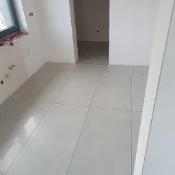 Wnętrze w trakcie remontu: świeżo ułożone jasnoszare płytki podłogowe z ciemnymi fugami, widoczne ślady po instalacjach na ścianie, otwór drzwiowy do innego pomieszczenia.