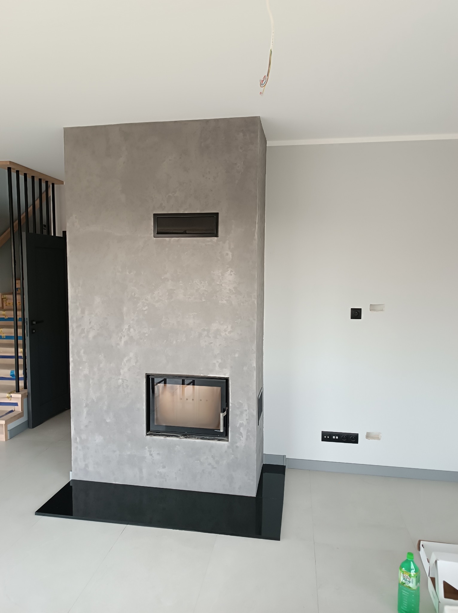 Nowoczesny kominek obudowany szarym tynkiem dekoracyjnym w surowym, minimalistycznym wnętrzu z białymi ścianami i podłogą, widoczne gniazdka elektryczne bez ramek i schody z czarną balustradą.