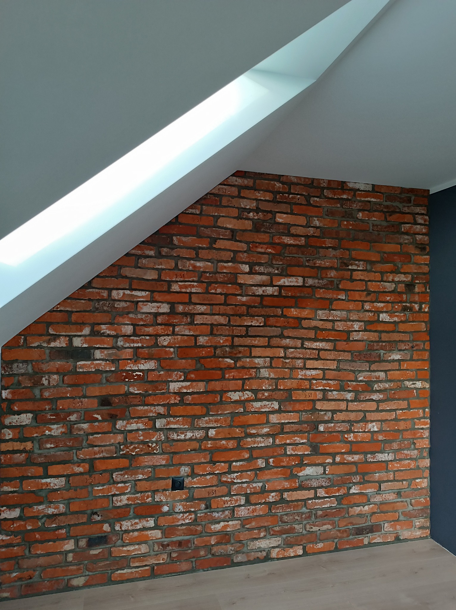 Ściana z czerwonej cegły z widoczną instalacją elektryczną, biały sufit skośny i fragment ściany pomalowanej na ciemny kolor.