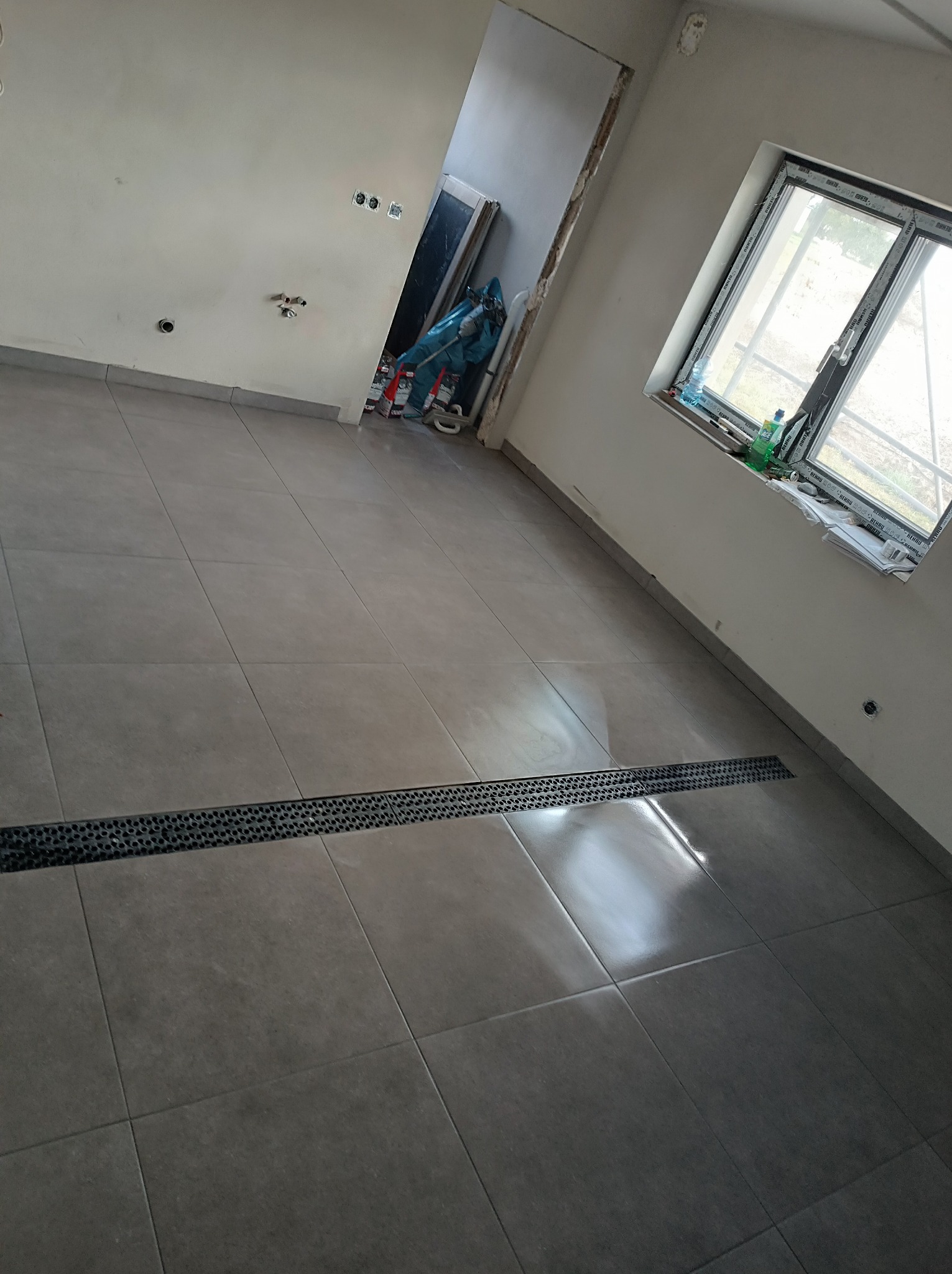 Szare płytki podłogowe z liniowym odpływem w nowym pomieszczeniu z oknem i widocznymi instalacjami elektrycznymi i wodnymi na ścianach.