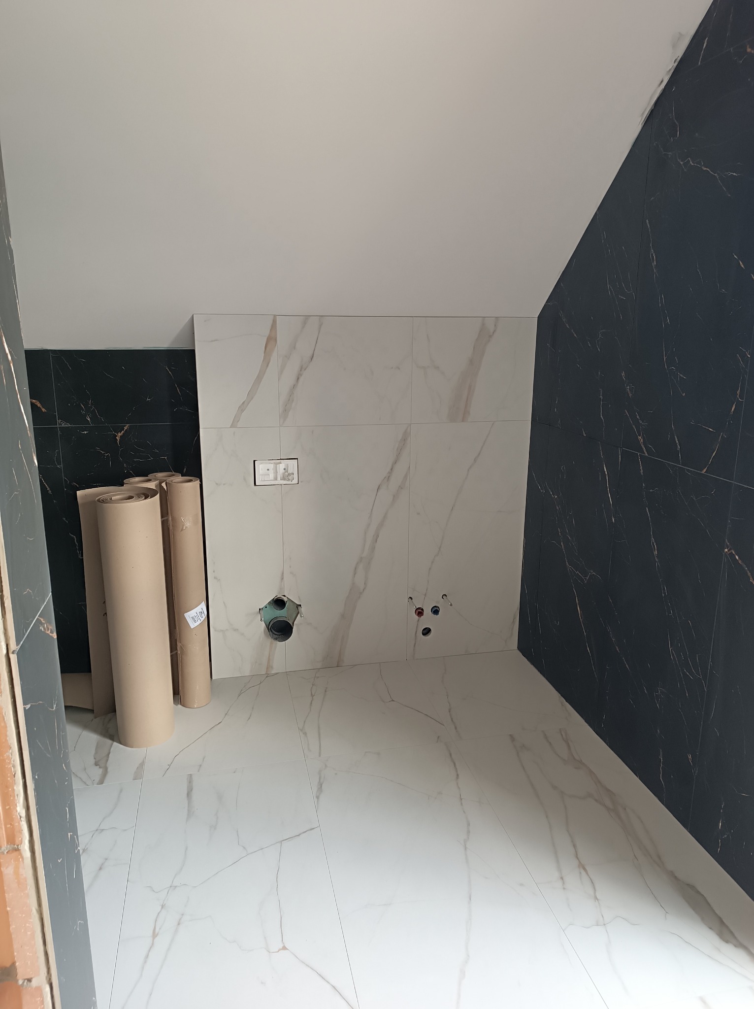Wnętrze łazienki w trakcie wykańczania: białe płytki imitujące marmur na podłodze i ścianie, czarne płytki na ścianie, rury kanalizacyjne i zwinięte rolki zabezpieczające podłogę.
