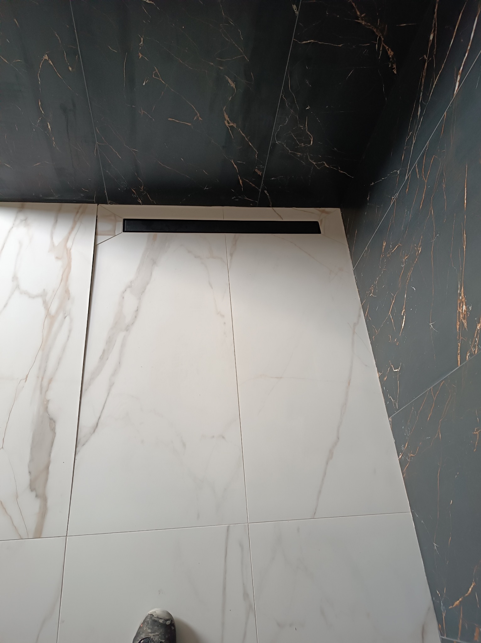Ułożone białe płytki podłogowe z marmurowym wzorem, czarny odpływ liniowy, fragment ciemnej ściany z wzorem żyłkowania i widoczny but roboczy w rogu kadru.