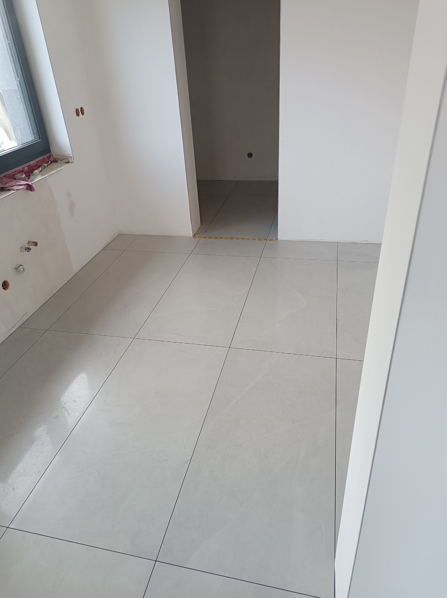 Wnętrze w trakcie remontu: świeżo ułożone jasnoszare płytki podłogowe z ciemnymi fugami, widoczne ślady po instalacjach na ścianie, otwór drzwiowy do innego pomieszczenia.