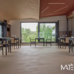 Wnętrze restauracji z jasną, jednolitą podłogą imitującą beton, ciemnymi stolikami nakrytymi szkłem i dużymi oknami z widokiem na zieleń.