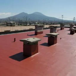 Czerwony dach pokryty nową powłoką hydroizolacyjną, z kominami w rzędzie i widokiem na miasto oraz góry w tle.