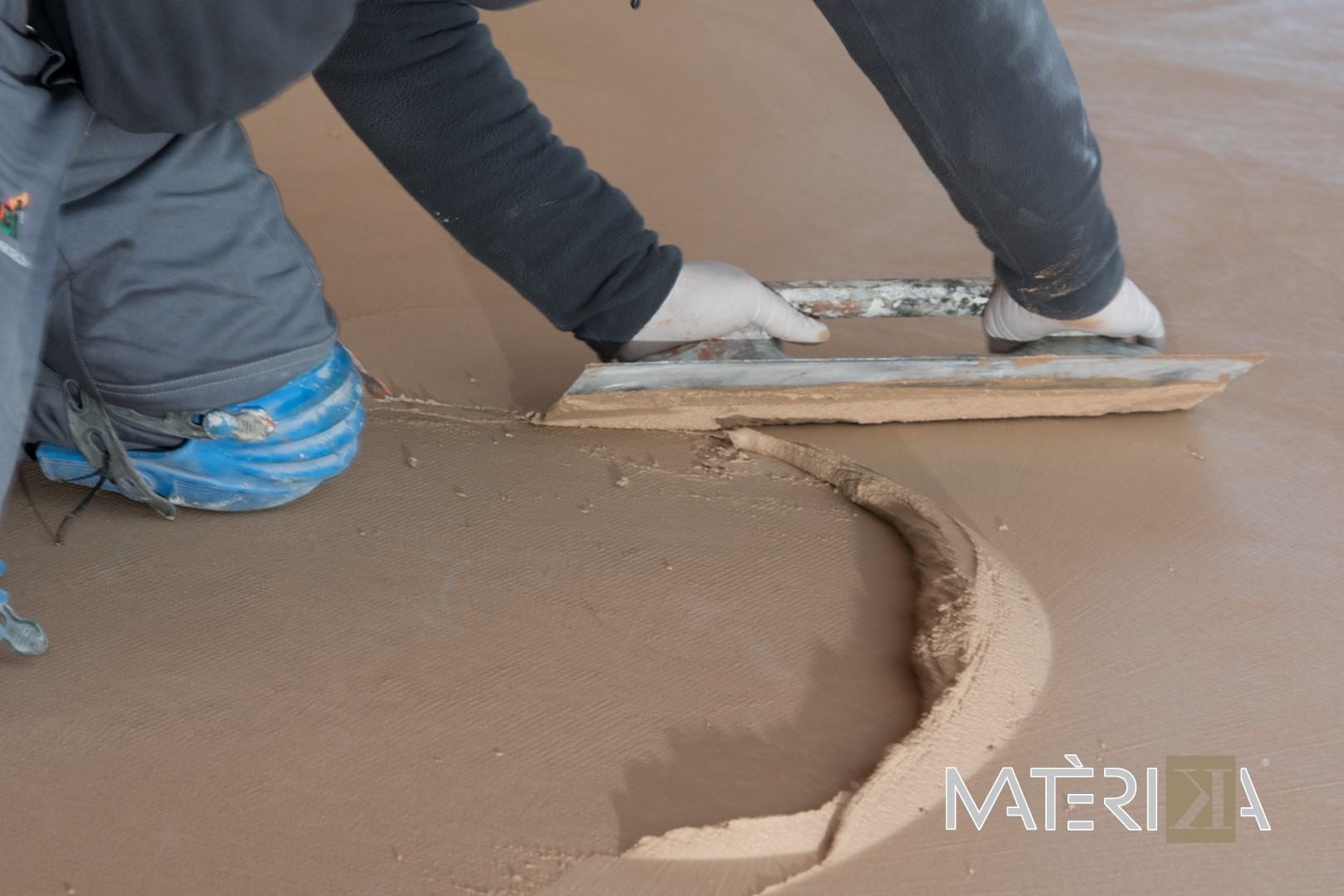 Aplikacja beżowej masy wyrównującej na podłodze przy użyciu pacy; widoczne ślady po narzędziu i fragment roboczych butów.