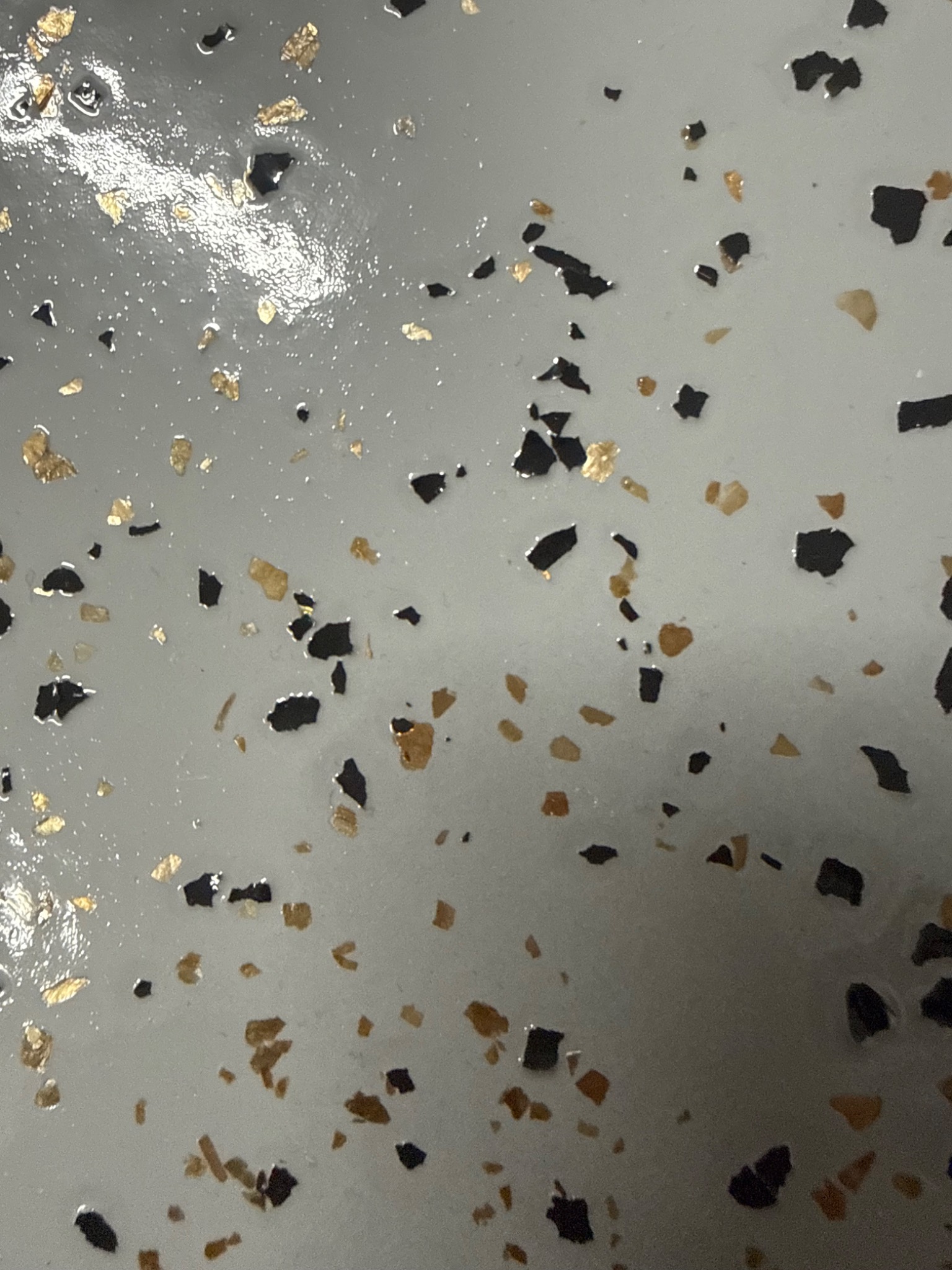 Błyszcząca, szara posadzka żywiczna z zatopionymi, nieregularnymi płatkami złota i czerni, tworzącymi unikalny wzór. Gładka powierzchnia z refleksami światła.