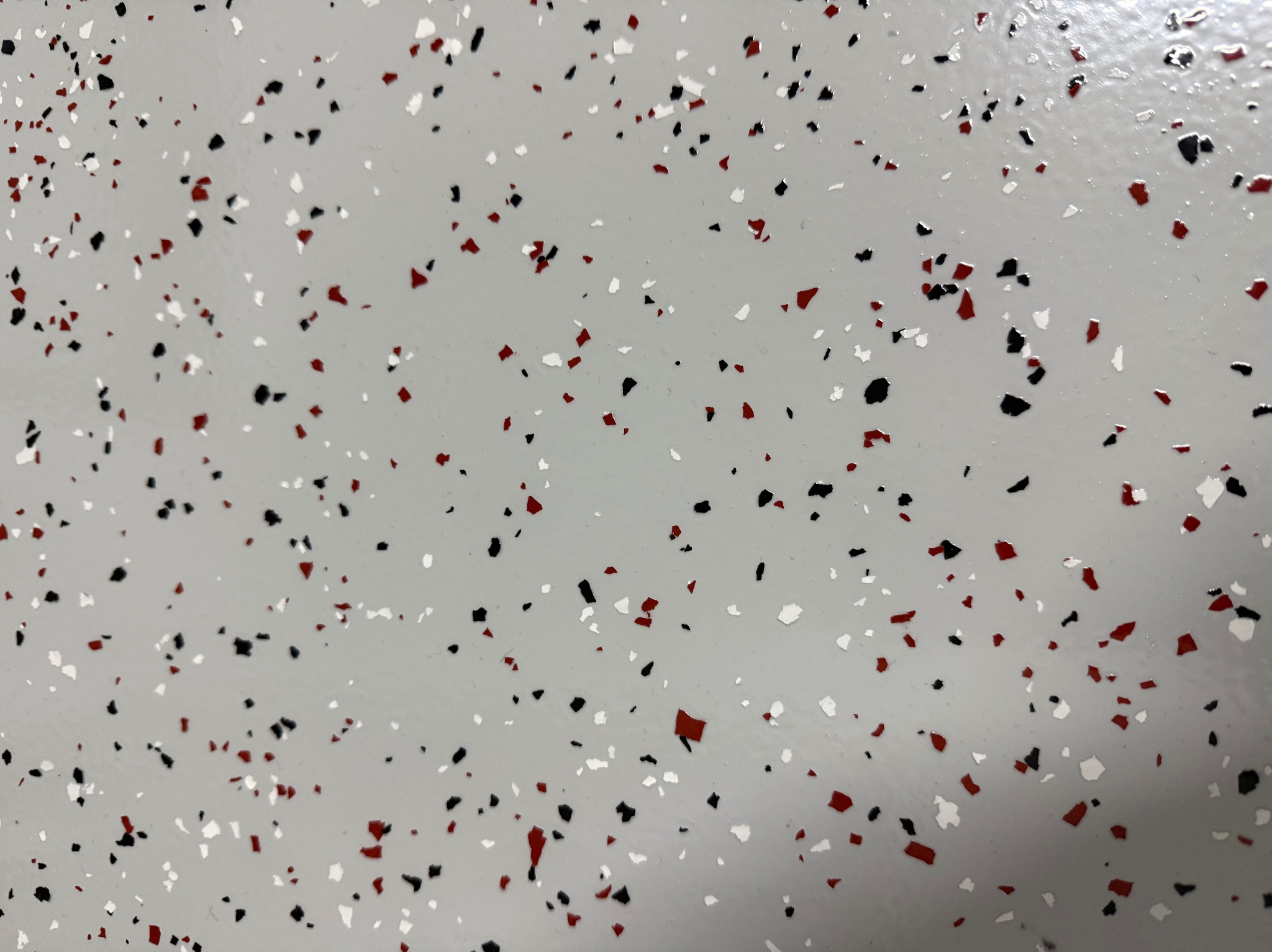 Gładka, jasnoszara posadzka żywiczna z zatopionymi, nieregularnymi płatkami w kolorach: białym, czarnym i czerwonym. Widok z góry, wypełniający kadr.