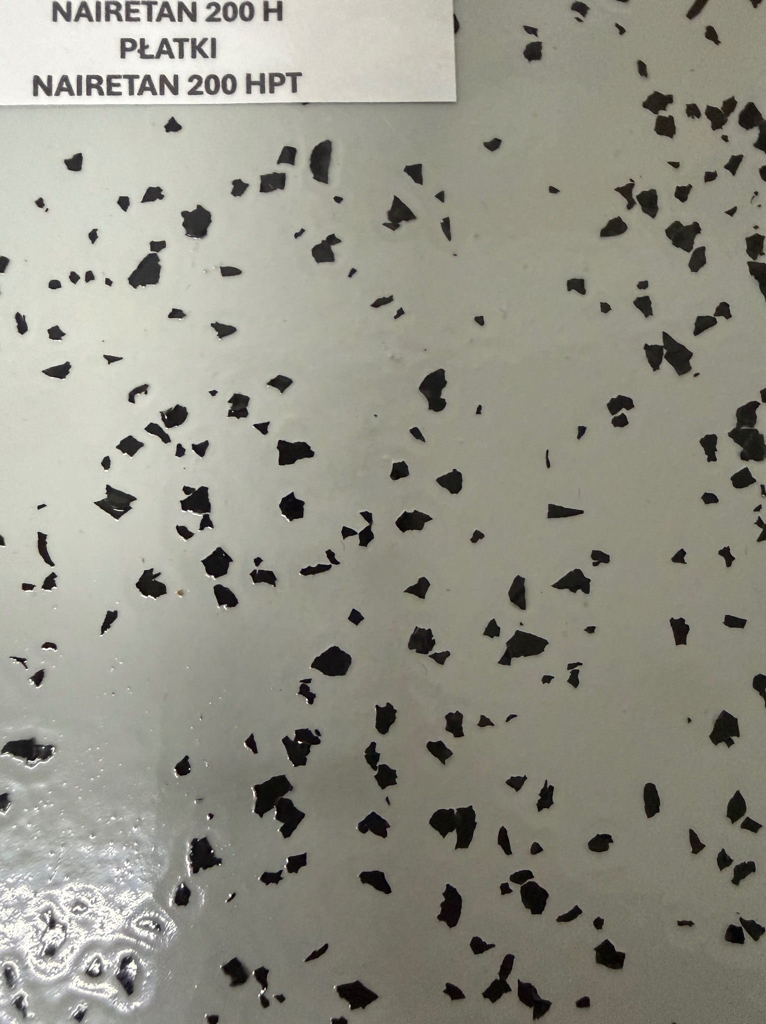 Szara, błyszcząca posadzka żywiczna z czarnymi płatkami dekoracyjnymi. W rogu widoczna etykieta z nazwą produktu: NAIRETRAN 200 H.