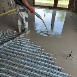 Wylewka podłogowa zalewana na ogrzewanie podłogowe, widoczny pracownik w czerwonych rękawicach i kaloszach, betonowa ściana w tle