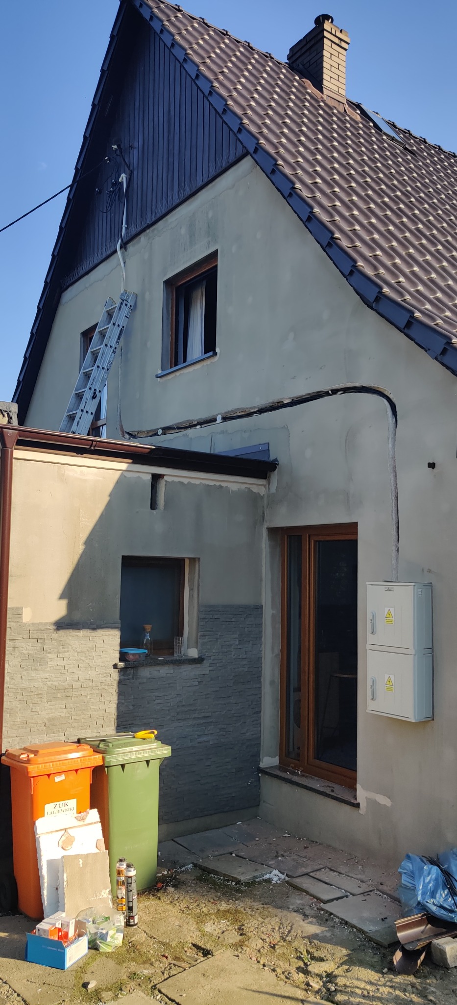 Zewnętrzna instalacja elektryczna na elewacji domu, widoczna puszka elektryczna, przewody w peszlu oraz drabina oparta o ścianę. W tle brązowy dach i niebieskie niebo.