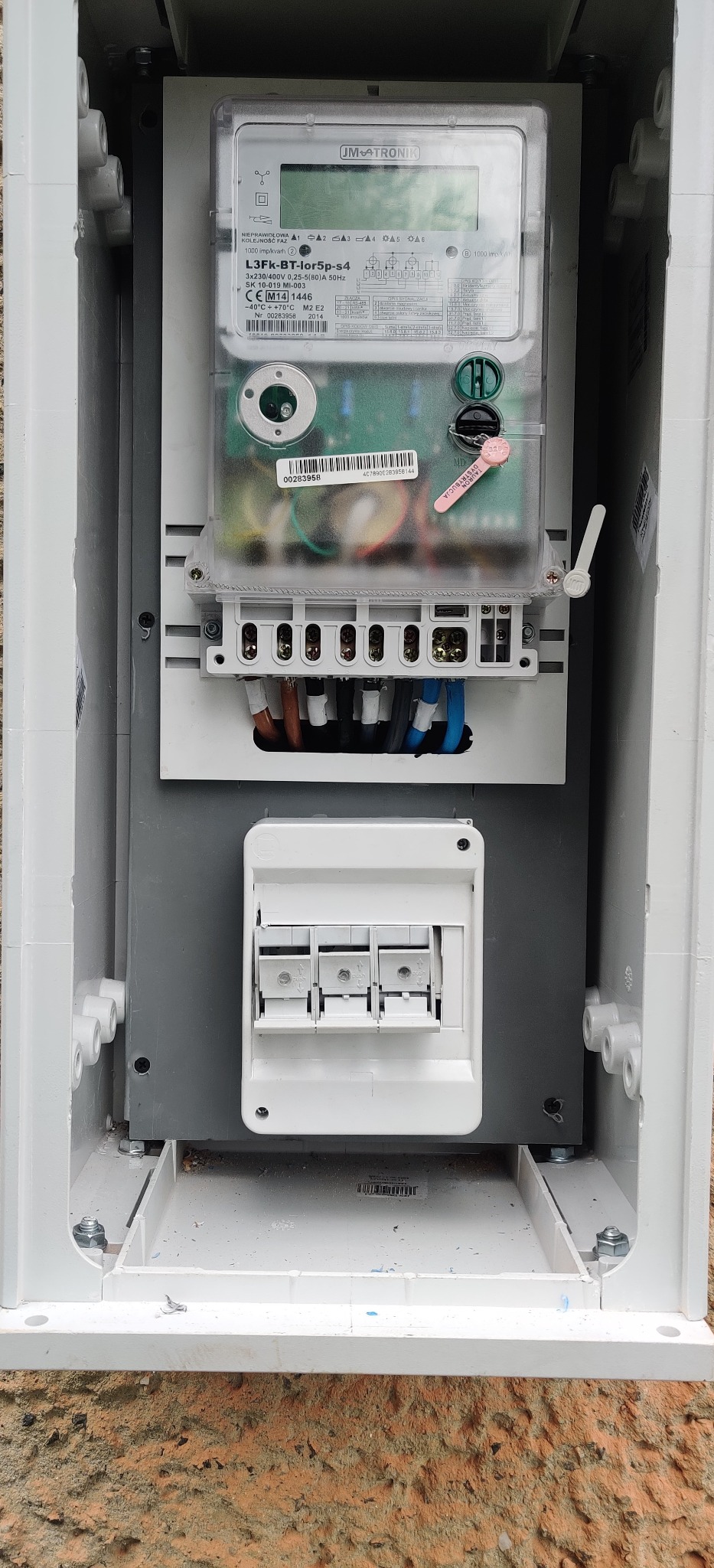 Zamknięta skrzynka elektryczna z licznikiem JM TRONIK L3Fk-BT-lor5p-s4, widoczne przewody i bezpieczniki.