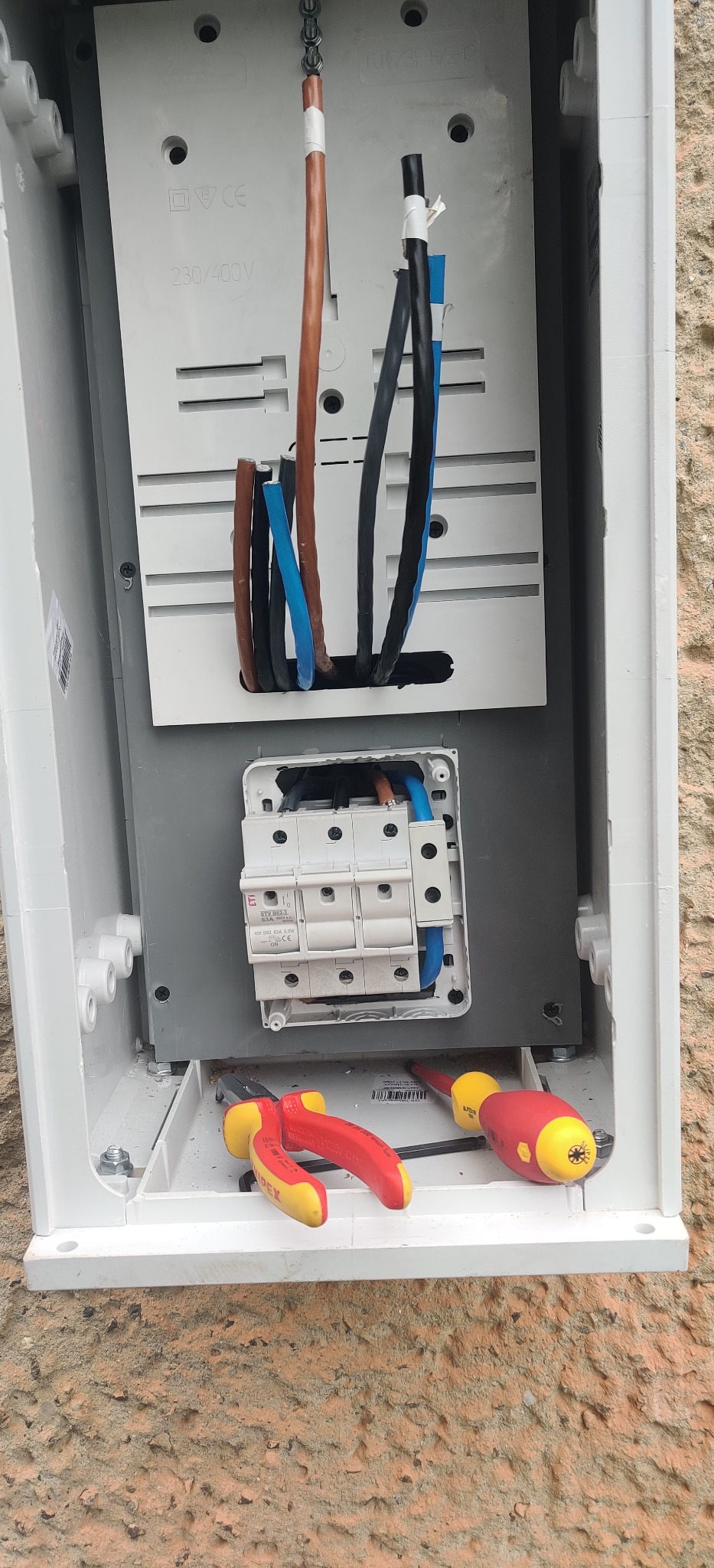 Otwarta skrzynka elektryczna z bezpiecznikami, widoczne przewody w izolacji brązowej, niebieskiej i czarnej, oraz narzędzia: kombinerki Knipex i śrubokręt z żółto-czerwoną rączką.