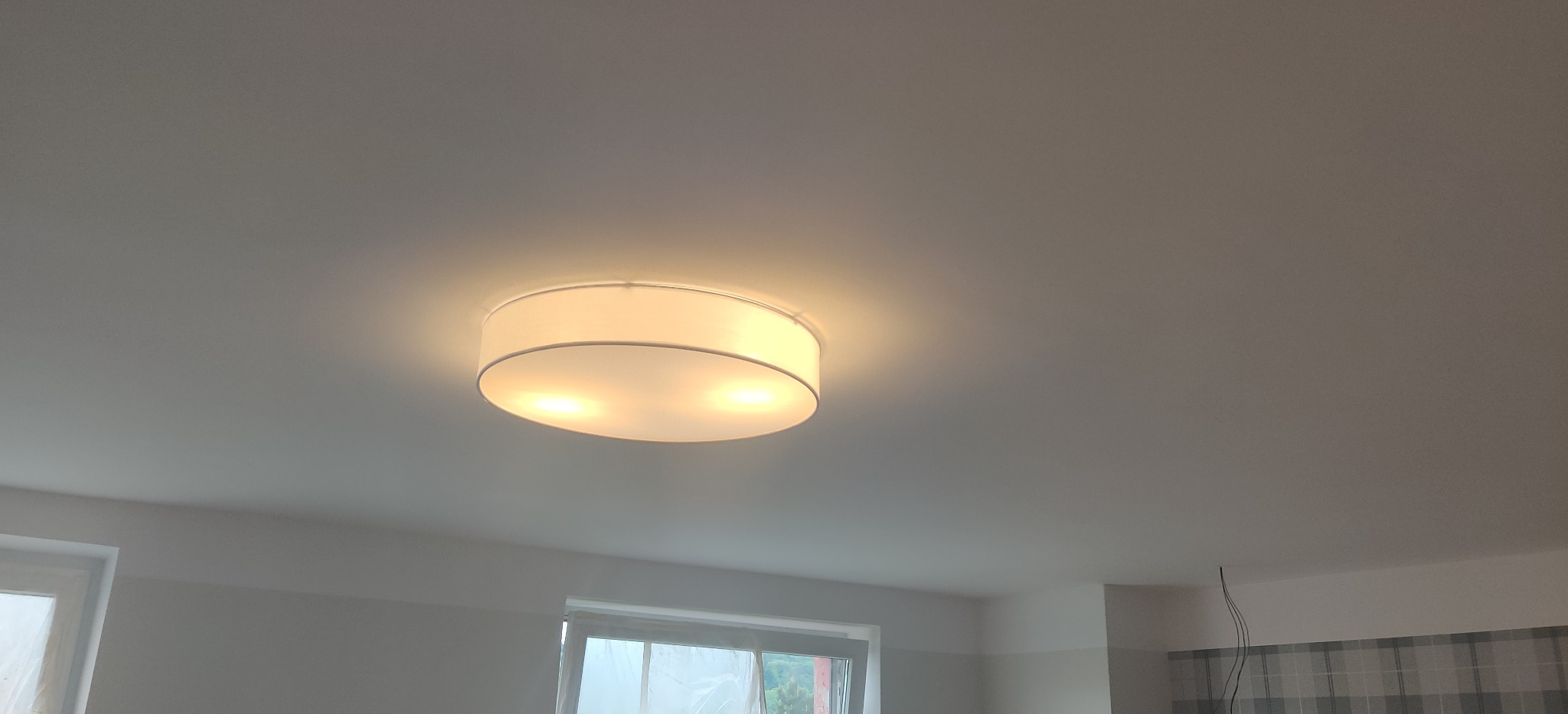 Zamontowana okrągła lampa sufitowa z białym abażurem, świecąca ciepłym światłem, widoczna na tle białego sufitu w nowym budownictwie.