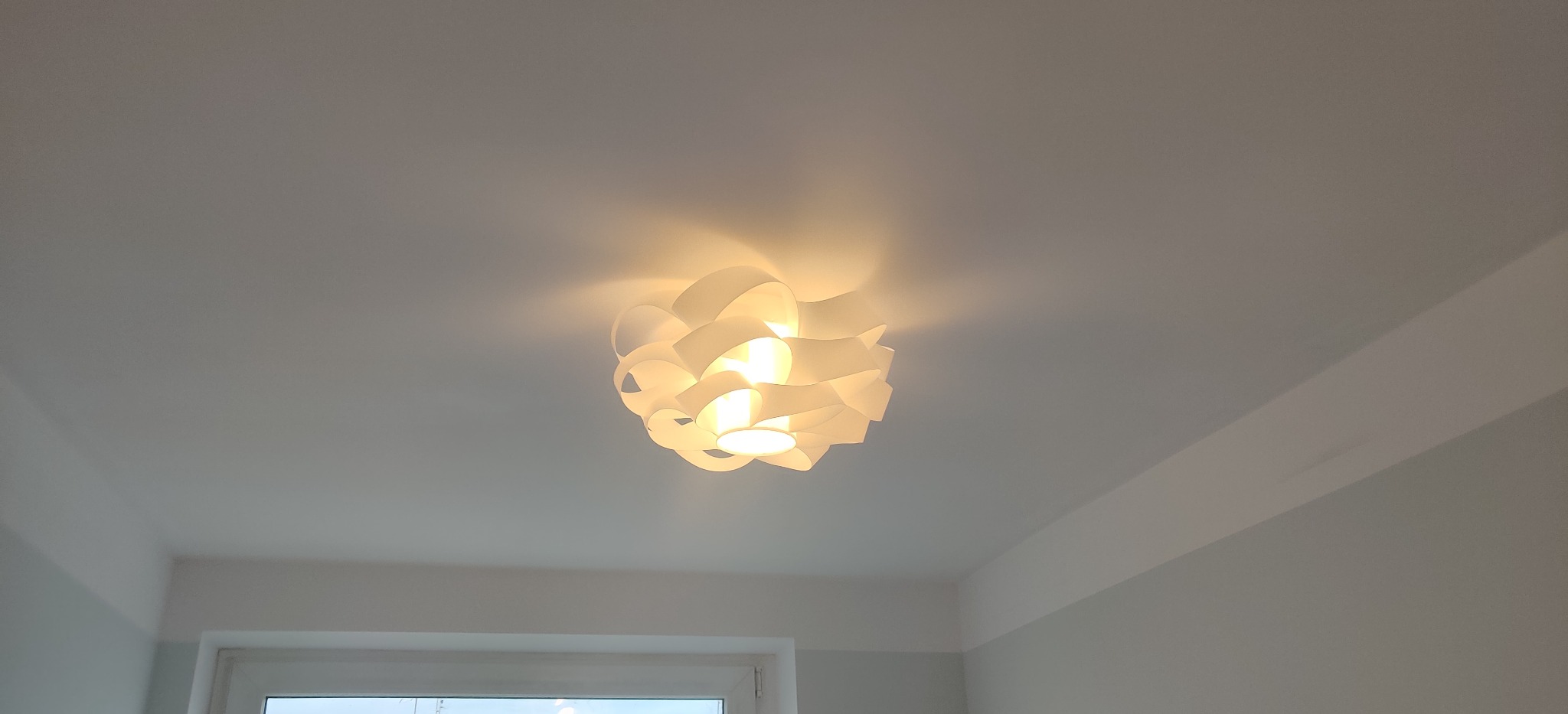 Biała lampa sufitowa z kloszem o falistym kształcie, oświetlająca białe wnętrze, widoczne okno w dolnej części kadru.