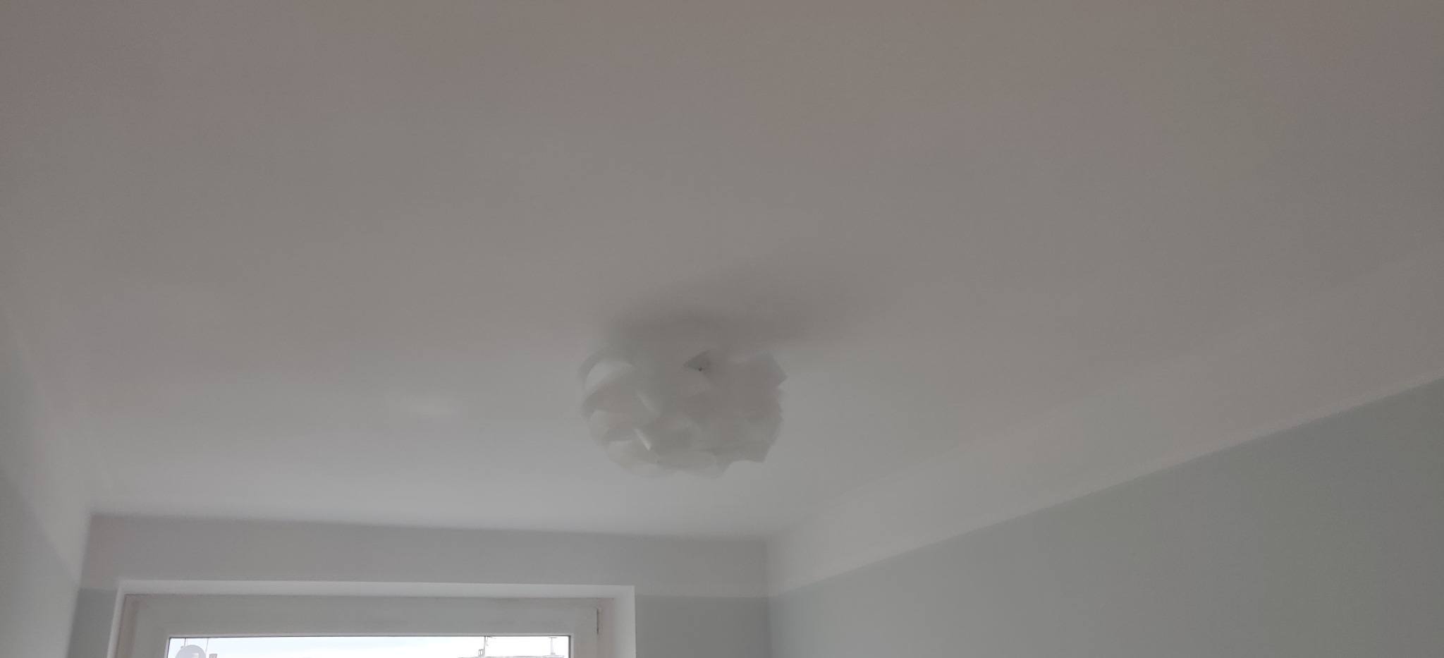 Biała, dekoracyjna lampa sufitowa w kształcie rozety, zamontowana na białym suficie w jasnym pomieszczeniu z widocznym fragmentem okna.