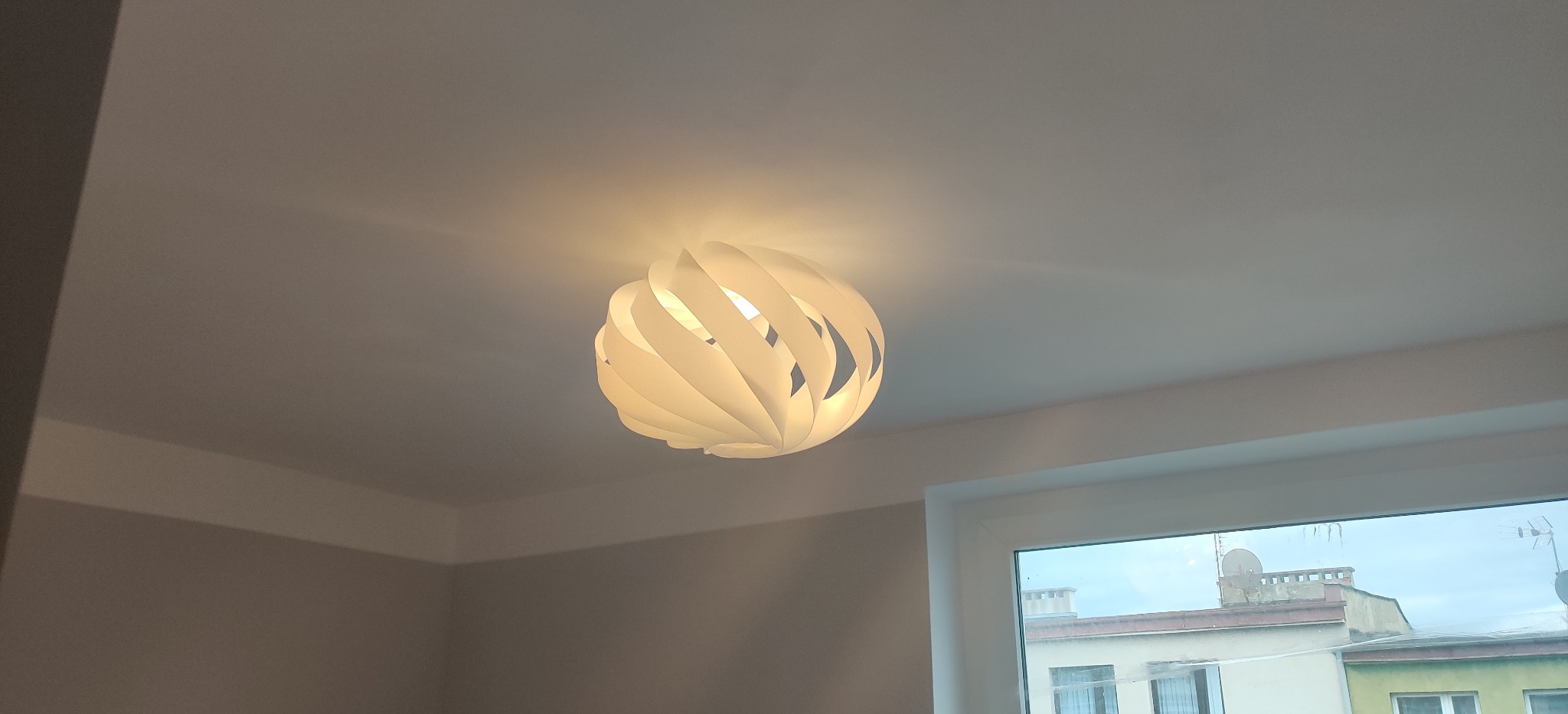 Zamontowana na suficie biała, nowoczesna lampa wisząca o kulistym kształcie z ażurowymi wycięciami, oświetlająca wnętrze, widok z okna na budynki w tle.