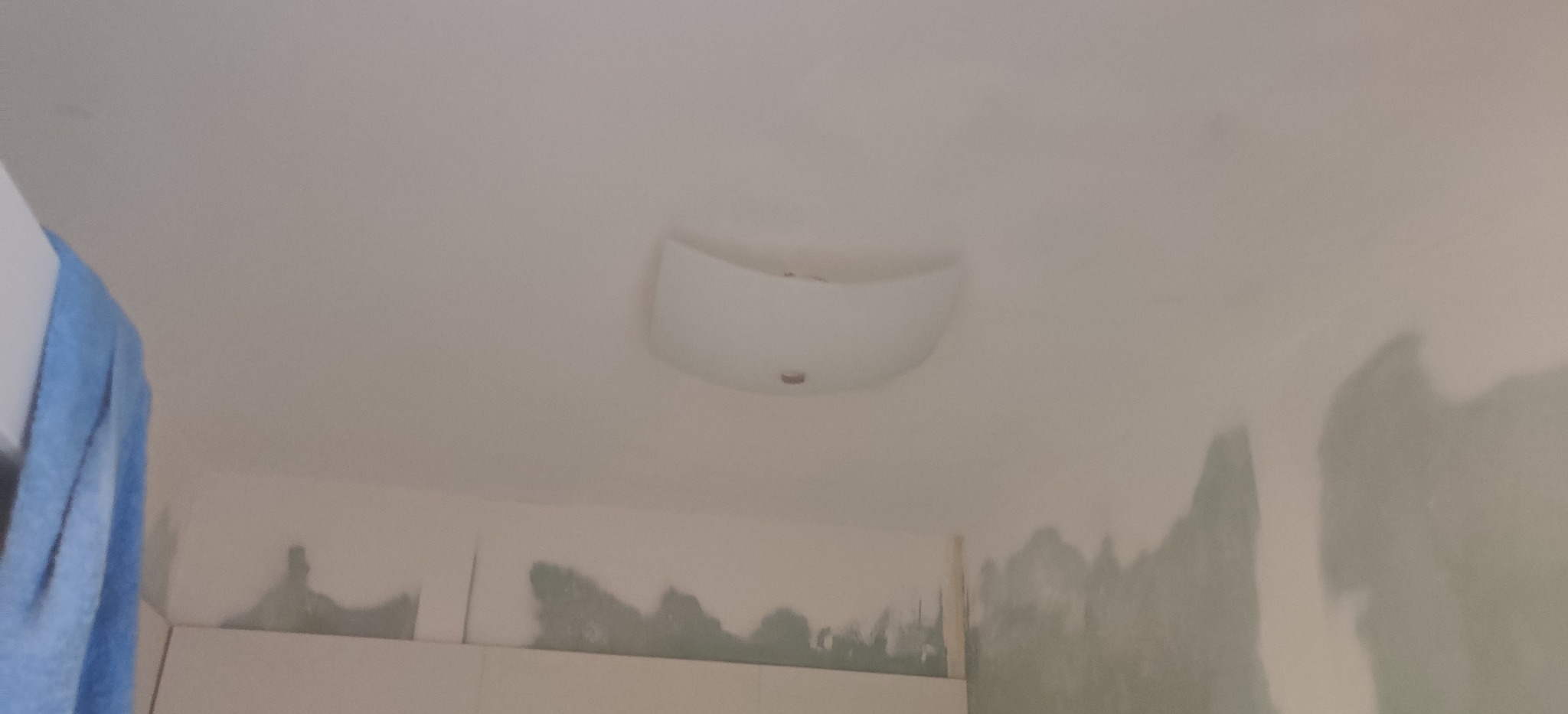 Zamontowana lampa sufitowa w łazience, widoczne ślady po malowaniu na ścianach, fragment niebieskiego ręcznika z lewej strony.