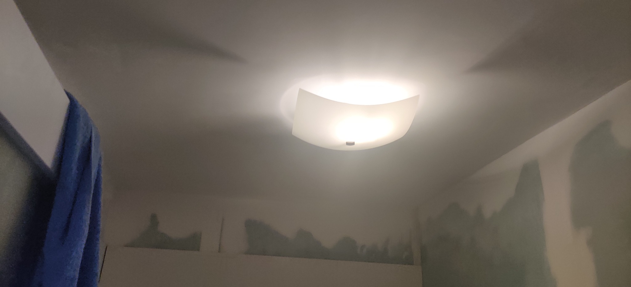 Lampa sufitowa z białym kloszem w łazience w trakcie remontu, widoczne ślady szpachlowania na ścianach, fragment niebieskiego ręcznika po lewej stronie.