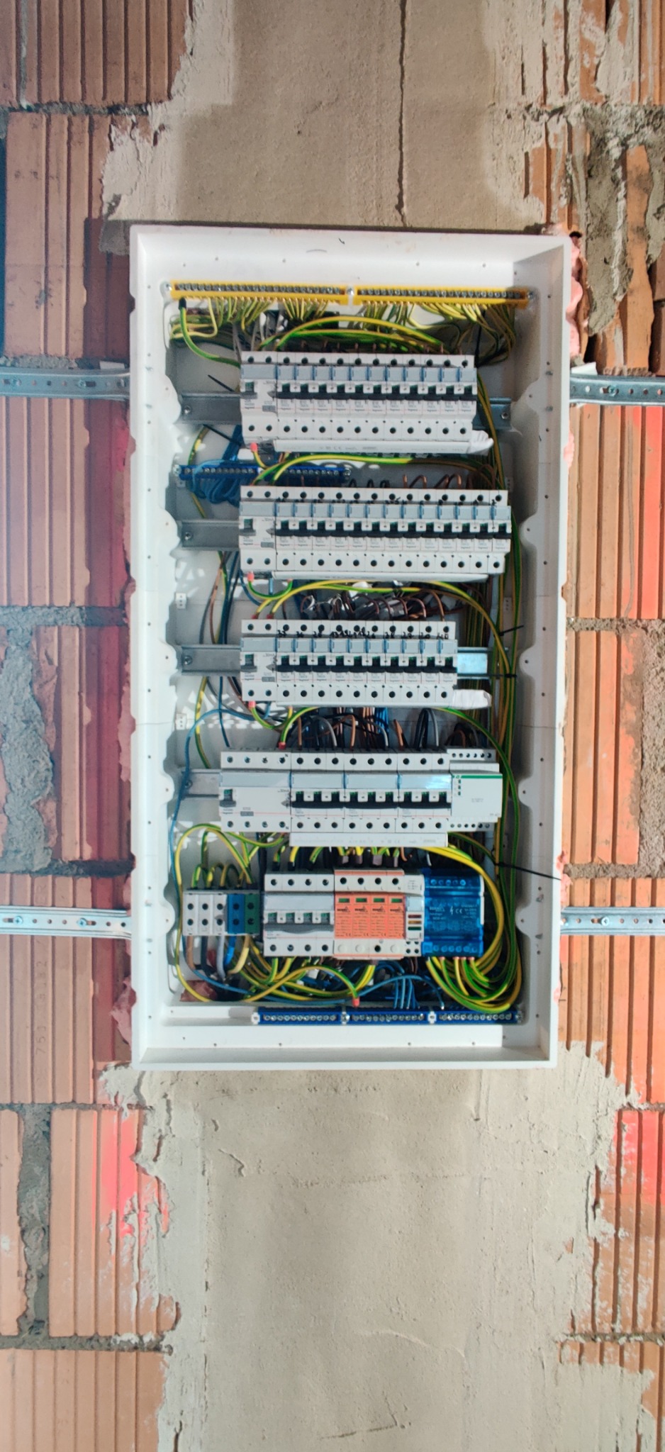 Otwarta skrzynka elektryczna z zabezpieczeniami różnicowoprądowymi i nadprądowymi, okablowaniem w kolorach żółto-zielonym, niebieskim i czarnym, zamontowana na ścianie z cegły i surowego tynku.