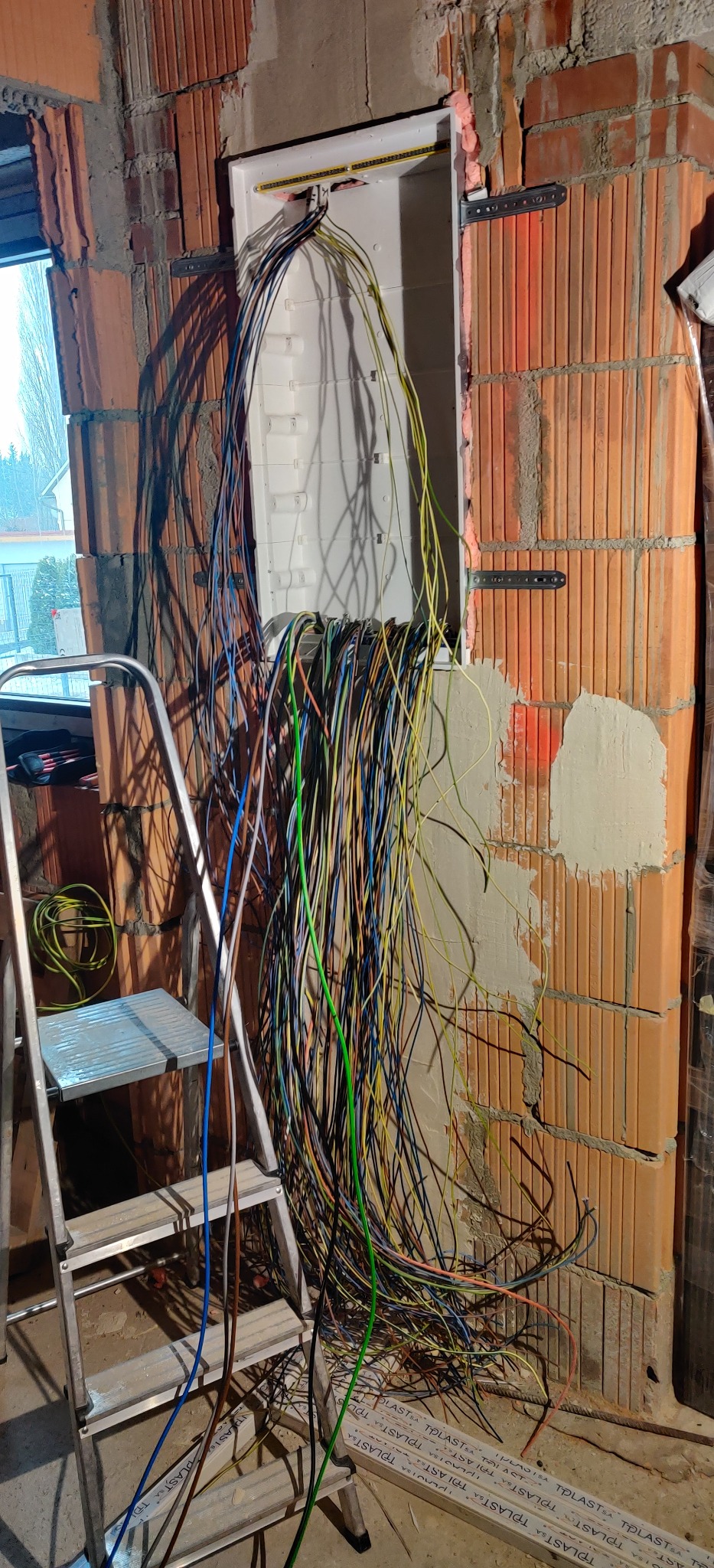 Widok na rozdzielnicę elektryczną w trakcie montażu w surowym stanie, z licznymi przewodami zwisającymi z puszki, drabina aluminiowa oparta o ścianę z cegły, widoczne ślady zaprawy i pianki...