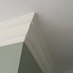 Szczegółowe ujęcie narożnika ściany i sufitu z listwą ozdobną, świeżo pomalowane na biało, z delikatnym odcieniem zieleni na ścianie.