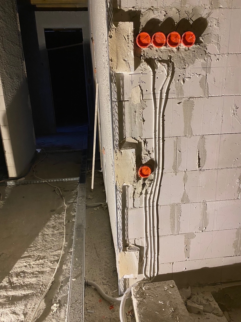 Surowa ściana z betonu komórkowego z widocznymi przewodami elektrycznymi w peszlach, pomarańczowe puszki instalacyjne pod gniazdka i włączniki, metalowy profil pod płytę gipsową przy ścianie, stan...