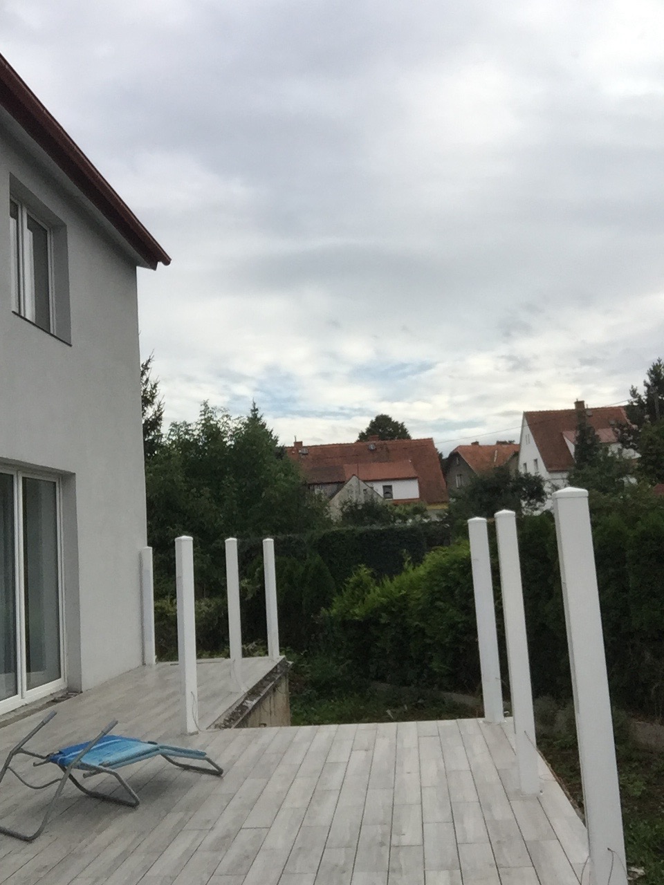 Szary taras z drewnopodobnych desek kompozytowych, białe słupki balustrady, widok na ogród i domy w tle, pochmurne niebo.