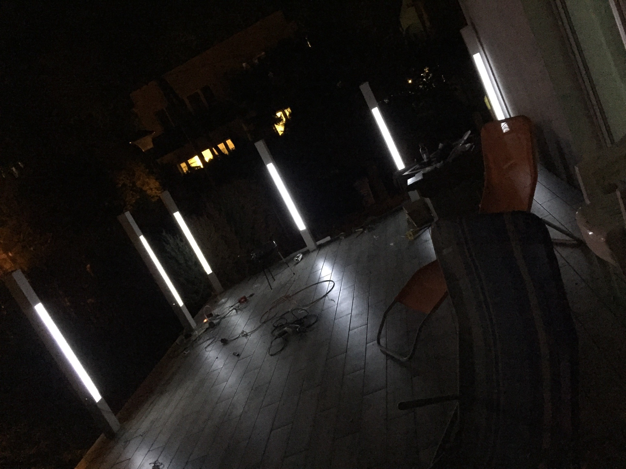 Nocny widok tarasu z podświetlanymi słupkami, kable i narzędzia na płytkach, krzesło w rogu, oświetlony budynek w tle.