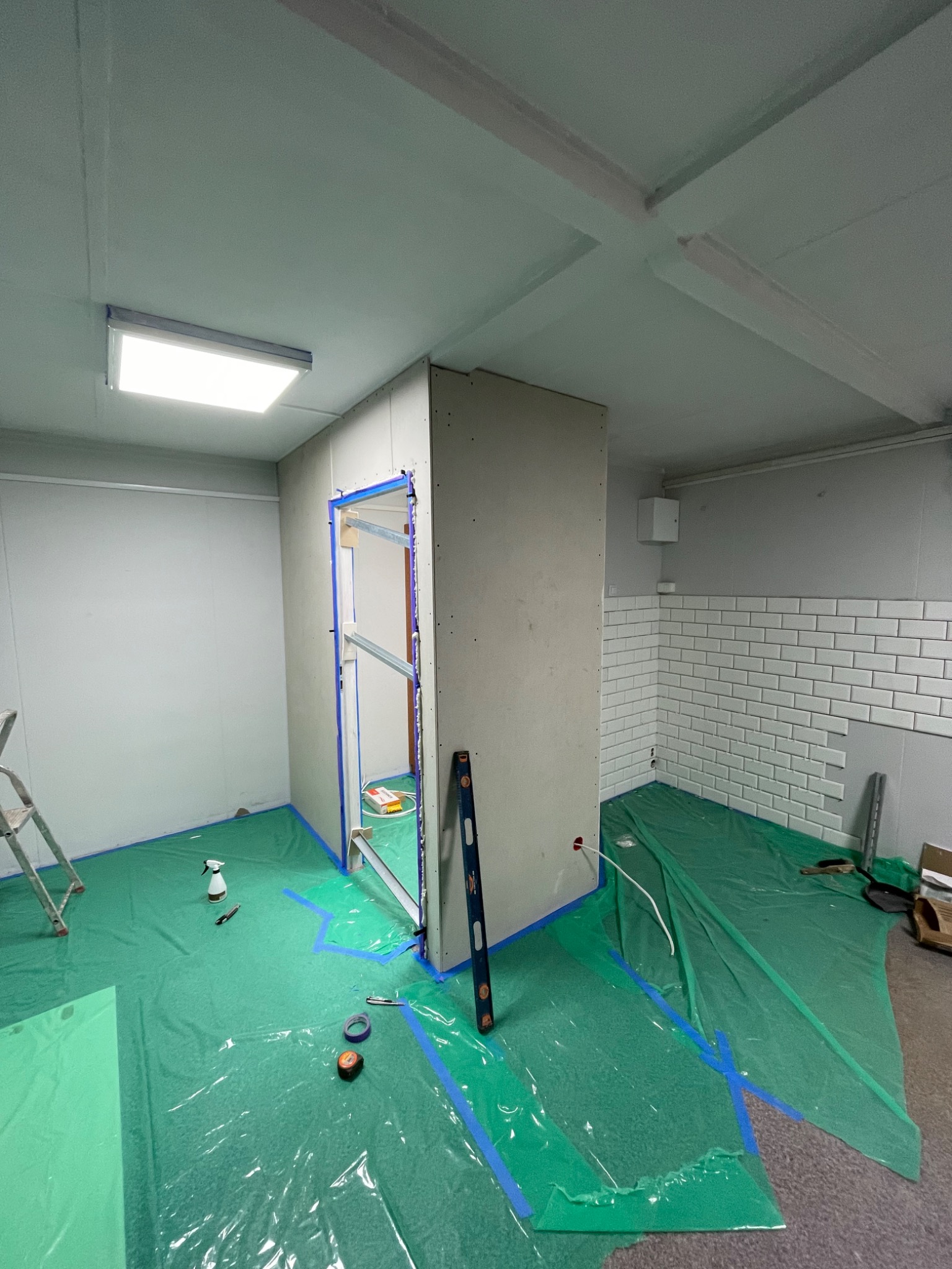 Wnętrze w trakcie prac: montaż ścianki działowej z płyt karton-gips, widoczna oklejona taśmą ościeżnica drzwiowa, poziomica przy ścianie, zabezpieczona folią podłoga i fragment ściany z białymi...