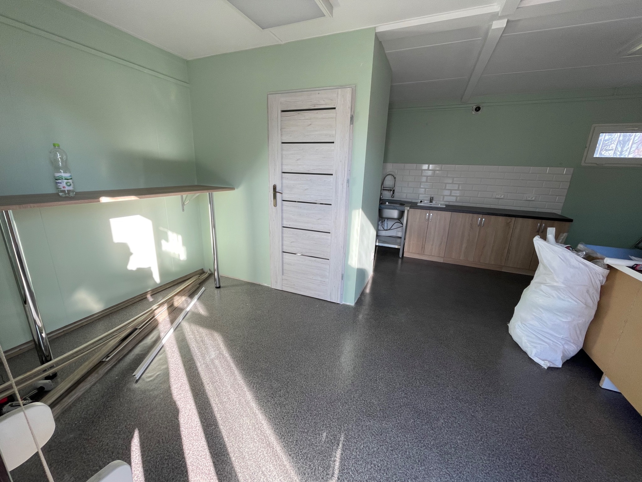 Wnętrze w trakcie remontu: świeżo pomalowane ściany w odcieniach zieleni, widoczna część kuchni z zabudową, drzwi wewnętrzne z poziomymi frezami, podłoga pokryta szarą wykładziną, elementy...